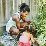 Smiling Mother And Young Daughter Tending To Plant Beds In Backyard Garden