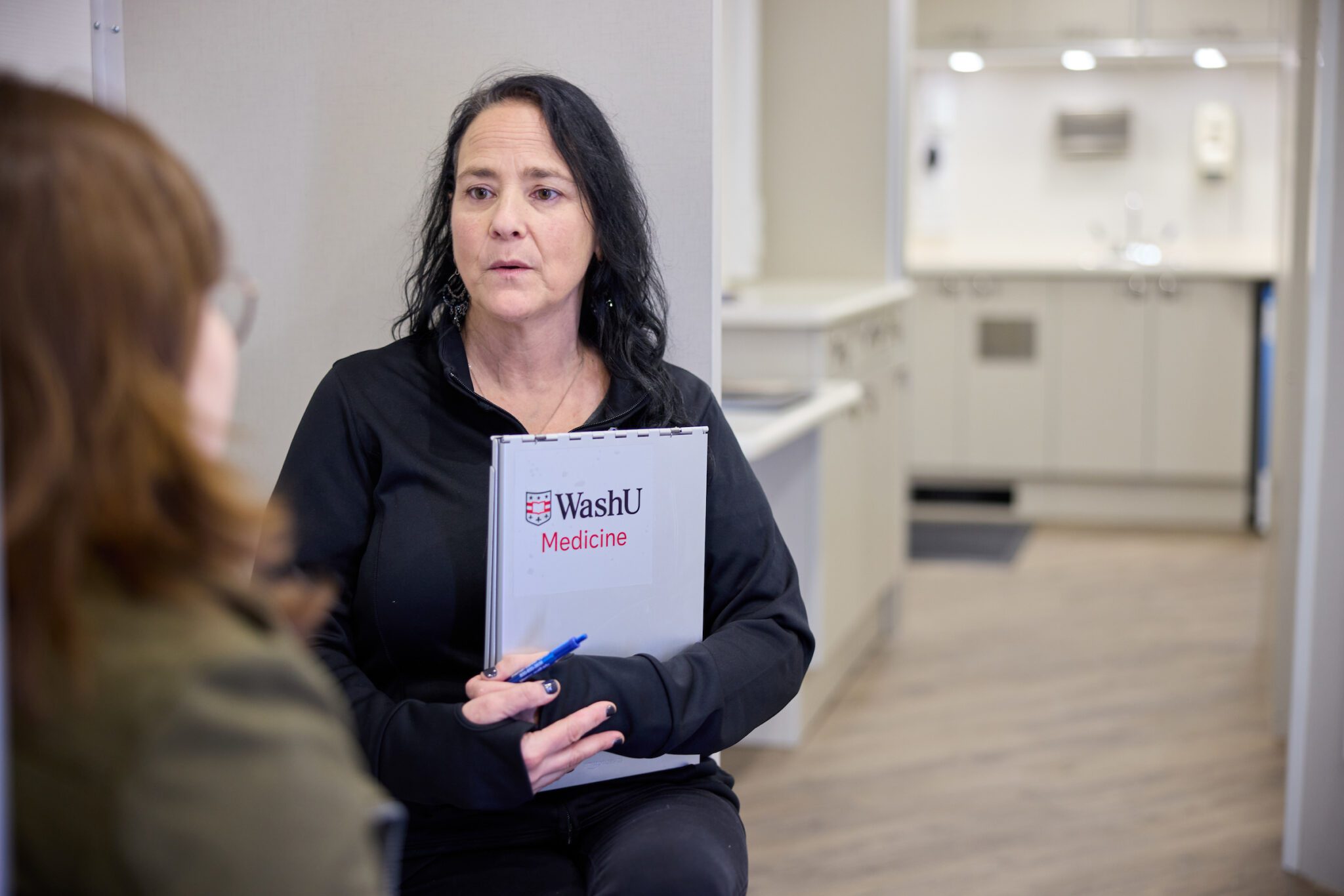 WashU expert on PECaD community van holds clipboard and consults with a patient inside white medical room.