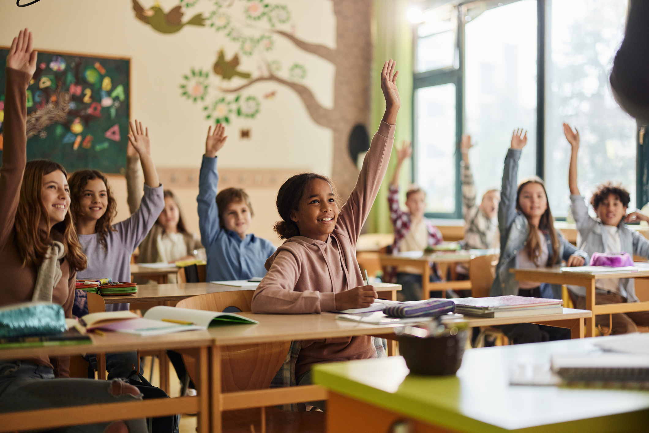 Happy Elementary Students Raising Their Hands On A Class At School.